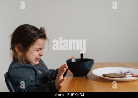 Une fillette malheureuse mange de la soupe dans un bol noir avec du pain et de l'oignon.Photo de style de vie d'un enfant dans la cuisine ayant un repas, hurlant enfant.Mangeur difficile Banque D'Images