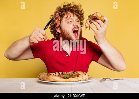 Homme coiffé à barbe rouge mangeant une délicieuse pizza italienne isolée sur fond jaune de studio.Concept de journée mondiale de la pizza. Banque D'Images