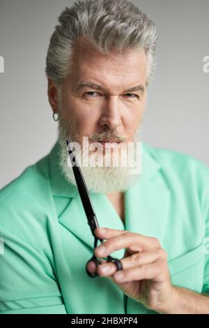 Portrait d'un homme d'âge moyen brutal avec une barbe regardant l'appareil photo, posant avec des ciseaux de barbier pointus sur fond blanc Banque D'Images