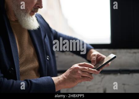 Prise de vue courte d'un homme d'âge moyen barbu en tenue professionnelle décontractée à l'aide de son smartphone, assis près d'une fenêtre dans un intérieur loft moderne Banque D'Images