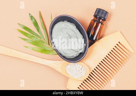 Masque cosmétique fait maison en argile avec huile essentielle à côté du peigne en bois et de la feuille verte sur fond marron, vue du dessus Banque D'Images