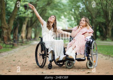 Agréables jeunes femmes handicapées souriant et s'amusant pendant la marche d'été au parc de la ville verte.Les femmes en fauteuil roulant profitent de journées chaudes à l'air frais. Banque D'Images