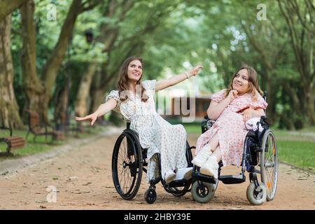 Agréables jeunes femmes handicapées souriant et s'amusant pendant la marche d'été au parc de la ville verte.Les femmes en fauteuil roulant profitent de journées chaudes à l'air frais. Banque D'Images