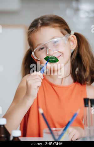 Mignonne souriante petite fille faisant un projet scientifique, tenant une feuille dans des pincettes.Elle porte des lunettes de protection, debout derrière une table. Banque D'Images