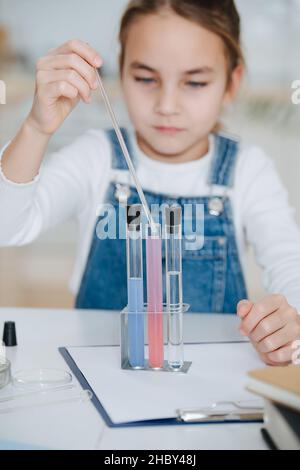 Petite fille floue faisant à la maison projet scientifique, remplissant une pipette avec le liquide de la fiole.Derrière une table. Banque D'Images