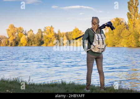 Femme tenant un trépied avec un appareil photo et cherchant la composition en automne nature près du lac.Femme photographe dans la nature Banque D'Images