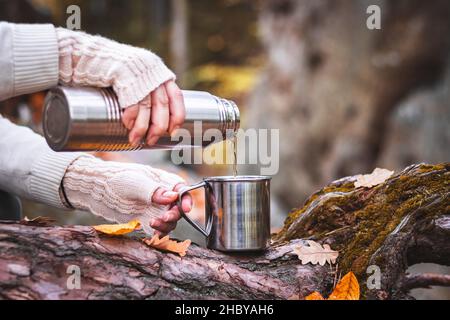 Une femme avec un gant tricoté verse une boisson chaude dans une tasse de thermos.Rafraîchissements pendant la randonnée. Banque D'Images