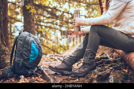 Femme en voyage avec sac à dos et pull buvant un café chaud d'une tasse et assis sur un arbre dans la forêt d'automne sauvage.Détente en randonnée. Banque D'Images