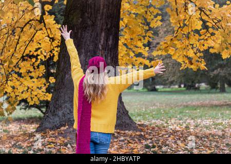 Femme heureuse profitant de la saison d'automne dans le parc public.Mannequin de mode portant un chandail tricoté jaune, un foulard en laine, un chapeau rouge et un Jean. Banque D'Images