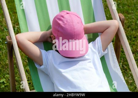 Jeune fille anonyme d'âge scolaire élémentaire, enfant dormant, se reposant, prenant une sieste sur le lit de soleil à l'extérieur avec une casquette de baseball sur son visage, o Banque D'Images