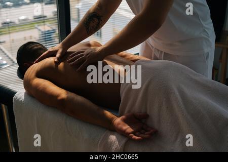 Coupe courte de masseuse masculine méconnue avec de fortes mains tatouées massant le dos et les épaules d'un sportif musclé allongé sur l'estomac Banque D'Images