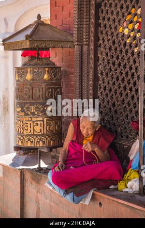 Une religieuse bouddhiste âgée est assise près d'une grande roue de prière de style tibétain près de la Boudhanath Stupa, Katmandou, Népal. Banque D'Images