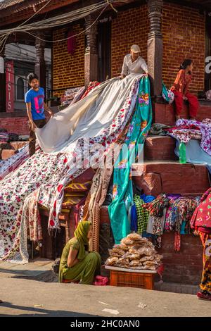 Deux hommes qui ont posé du tissu sur les marches d'un temple hindou à exposer pour la vente dans la rue de Katmandou, au Népal.Une femme vend des collations à l'avant. Banque D'Images