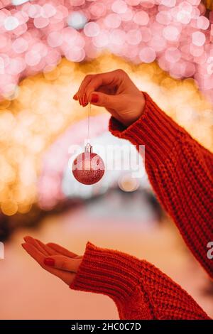 Les mains des femmes dans un chandail rouge tricoté tenant une boule de Noël étincelante sur un fond de lumières dorées festives Banque D'Images