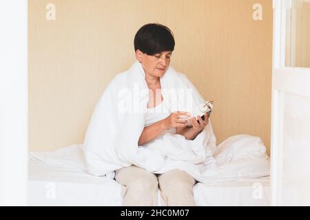 Une femme d'âge moyen est assise au lit avec un réveil entre ses mains.Réveil du matin, troubles du sommeil insomnie Banque D'Images