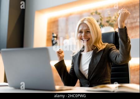 Confiante heureuse joyeuse femme d'affaires mûre, boursière, assis à table dans le bureau moderne, crient et se réjouit de la réussite, de la victoire ou du bon profit, regarde l'ordinateur portable, des gestes avec ses poings, sourire Banque D'Images