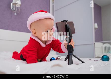 Un bébé garçon vêtu d'un costume de père Noël s'assoit sur le lit et regarde le téléphone malade.Vœux de Noël par appel vidéo Banque D'Images