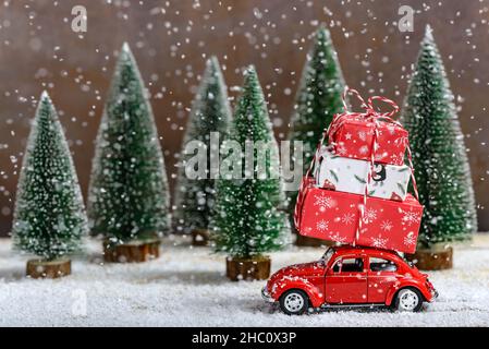 Voiture de jouet rouge avec un cadeau de Noël sur le toit à côté des sapins pendant qu'il neige sur une table en bois.Concept de célébration de Noël et du nouvel an. Banque D'Images