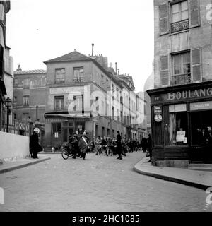 Une procession en 1945 à Paris Montmartre vient de la direction de Sacre-Cœur en descendant la rue Norvins et en tournant à gauche sur la place Jean-Baptiste Clément.Le meneur est un homme âgé portant un chapeau Napoléon, un manteau et des bottes d'équitation.Cigare en bouche, une épée se fait miroiter sur son côté gauche.Il semble avoir perdu son bras gauche.Derrière lui, en mars, il y a un groupe de garçons qui battent des tambours.Un homme de la Marine américaine s'est enrôé montres d'un côté et Une femme montres et puis rejoint à l'avant du groupe.La silhouette du dôme du Sacré-cœur est en vue, tout comme le Consulat d’Auvergne et le Restaurant l’Ambassade. Banque D'Images