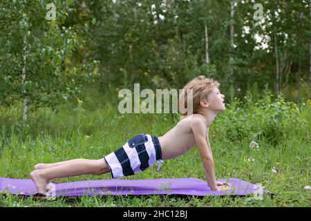 Petit garçon en short rayé fait de l'exercice sur le tapis de fitness dans le parc.Kid est engagé dans des exercices physiques dans la nature. Banque D'Images