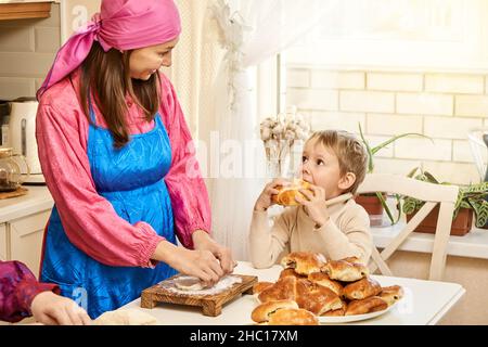 Une femme positive dans le costume national d'Udmurt pétrit la pâte à cuire des tartes avec la mère mûre tandis que le petit fils mange une tarte dans la cuisine spacieuse à la maison Banque D'Images