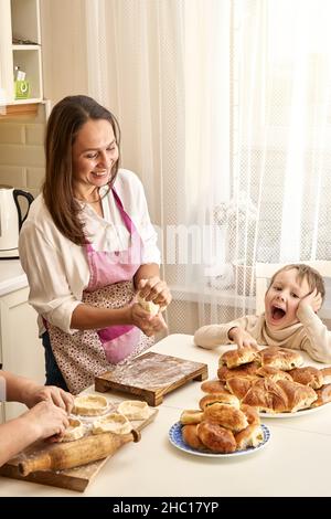 Une dame riant en tablier pétrit la pâte en préparant des tartes avec une mère âgée et un petit garçon à table blanche dans la cuisine légère à la maison Banque D'Images