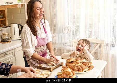 Une dame riant en tablier pétrit la pâte en préparant des tartes avec une mère âgée et un petit garçon à table blanche dans la cuisine légère à la maison Banque D'Images