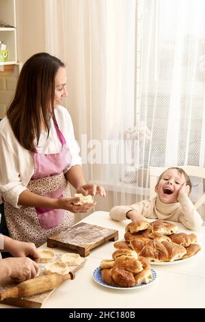 Une dame riant en tablier pétrit la pâte en préparant des tartes avec une mère âgée et un petit garçon à table blanche dans la cuisine légère à la maison Banque D'Images