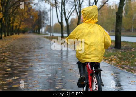 Un homme en imperméable jaune conduit le long d'une allée dans un parc d'automne.Garçon fait du vélo dans la ville sous la pluie.Vue arrière. Banque D'Images