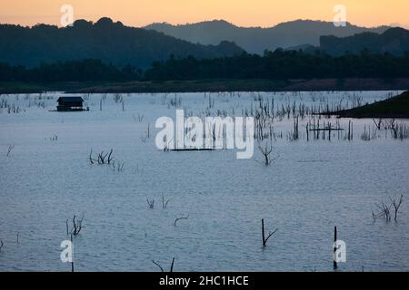 Le paysage d'un lac tranquille au crépuscule, une maison flottante, et le filet de radeau traditionnel sur un lac, des montagnes, et le ciel de coucher de soleil.Khao Laem, Thaïlande. Banque D'Images
