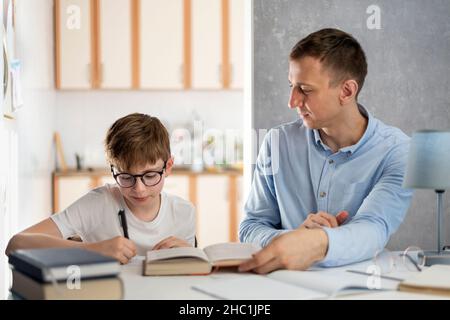 Le père et l'enfant font leurs devoirs.Le jeune professeur tuteur étudie avec le garçon.Garçon adolescent avec des lunettes écrit dans un carnet Banque D'Images