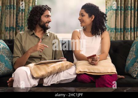 Jeune couple conversant pendant le shopping en ligne tout en étant assis sur le canapé à la maison Banque D'Images