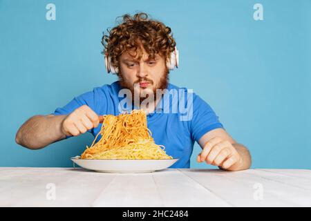 Jeune homme émotif barbu rouge et poiré goûtant une grande partie de nouilles, pâtes isolées sur fond bleu studio. Banque D'Images