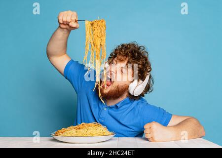 Drôle jeune poilu rouge-barbu homme mangeant une grande partie de nouilles, pâtes isolées sur fond bleu studio. Banque D'Images