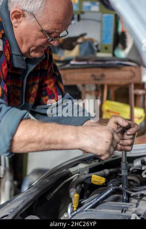 Portrait d'un mécanicien de voiture senior qui change les bougies d'allumage de sa vieille voiture dans le garage.Fixer les bougies d'allumage à l'aide d'un long tournevis spécial.Vieilles voitures. Banque D'Images