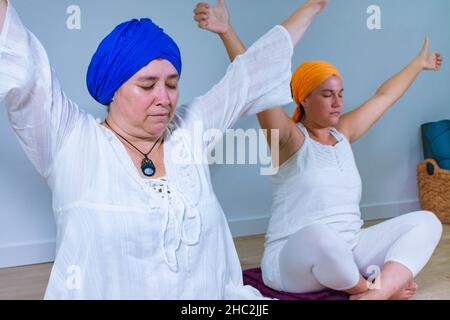 Deux femmes vêtues de blanc avec le turban pratiquant le yoga kundalini assis sur le sol avec les bras ouverts relevés Banque D'Images