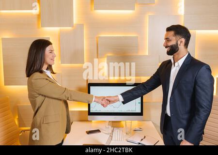 Joyeux jeune homme multiracial en costume se serrant les mains avec une femme caucasienne attrayante collègue.Concept d'ambiance de bureau Banque D'Images