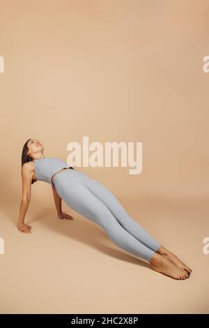 Brunette est en forme physique.Belle jeune femme en costume de yoga gris ajusté fait un exercice de planche inversée sur fond beige neutre, côté Banque D'Images