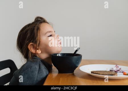 Une fillette malheureuse mange de la soupe dans un bol noir avec du pain et de l'oignon.Photo de style de vie d'un enfant dans la cuisine ayant un repas, hurlant enfant.Mangeur difficile Banque D'Images