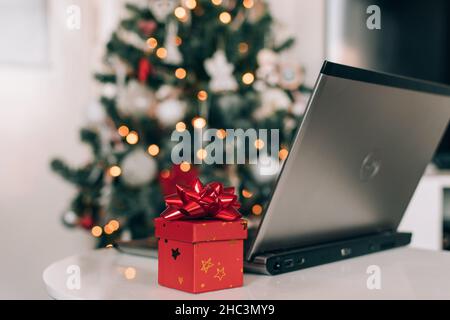 Petit cadeau rouge avec noeud cadeau à côté de l'ordinateur portable sur la table devant l'arbre de noël décoré Banque D'Images