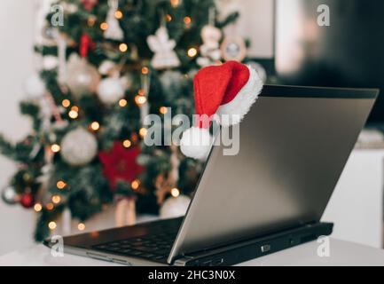 Chapeau de père noël sur le dessus de l'écran d'ordinateur portable devant un arbre de noël décoré avec le scintillement de bokeh Banque D'Images