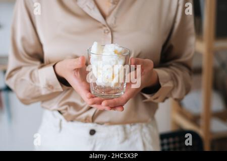 Gros plan d'un artisan féminin non reconnaissable tenant un verre transparent de cire de soja blanche et sèche pour créer un mélange de fabrication de bougies. Banque D'Images