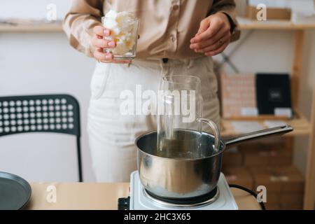 Gros plan de l'artisan féminin ajoutant de la cire de soja blanche et sèche dans un pot en verre dans un pot d'eau bouillante pour créer un mélange de fabrication de bougies. Banque D'Images