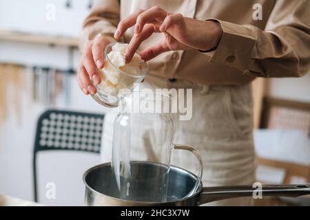 Gros plan de l'artisan féminin ajoutant de la cire de soja blanche et sèche dans un pot en verre dans un pot d'eau bouillante pour créer un mélange de fabrication de bougies. Banque D'Images