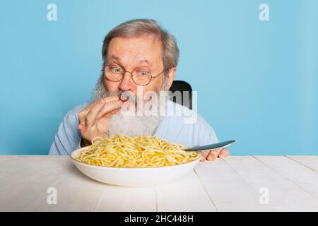 Emotionnel senior poilu gris-barbu homme mangeant une grande partie de nouilles, pâtes isolées sur fond bleu studio. Banque D'Images