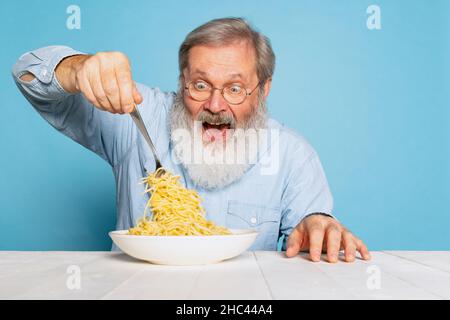 Homme à barbe grise et coiffée, qui mange une grande partie de nouilles, pâtes isolées sur fond de studio bleu. Banque D'Images