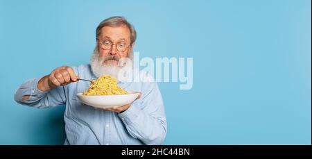 Surprise senior coiffé gris-barbu homme mangeant une grande partie de nouilles, pâtes isolées sur fond bleu studio. Banque D'Images