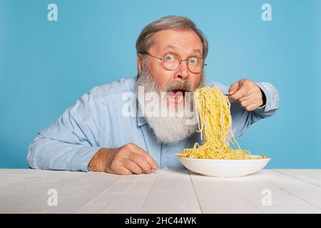 Choqué senior poilu gris-barbu homme mangeant une grande partie de nouilles, pâtes isolées sur fond bleu studio. Banque D'Images