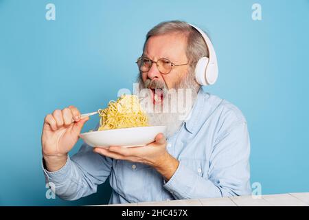 Emotionnel senior poilu gris-barbu homme mangeant une grande partie de nouilles, pâtes isolées sur fond bleu studio. Banque D'Images