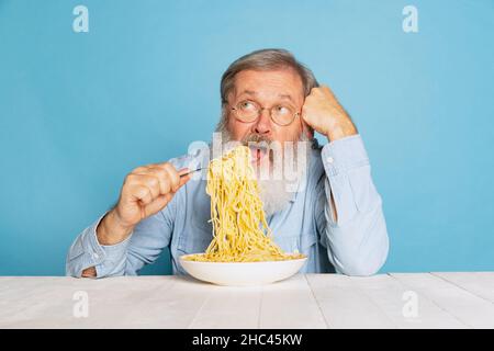 Emotionnel senior poilu gris-barbu homme mangeant une grande partie de nouilles, pâtes isolées sur fond bleu studio. Banque D'Images